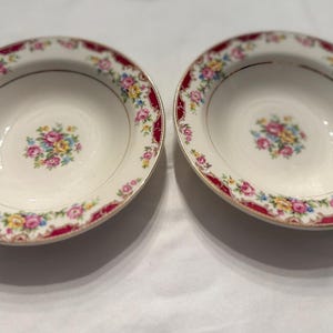 May include: Two oval-shaped bowls in off-white, featuring a floral pattern. The rims have a burgundy band and gold trim. Each bowl's centre displays a small bouquet of pink, yellow, and blue flowers. A classic tableware set.