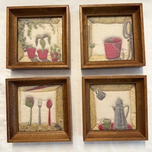 Könnte beinhalten: Vier gerahmte Wandteppich-Kunstwerke. Jeder quadratische Rahmen ist aus Holz gefertigt. Die Wandteppiche zeigen verschiedene Szenen: Topfpflanzen, einen roten Eimer, Besteck und eine Kaffeekanne mit einer Tasse. Die Wandteppiche haben einen neutralen Hintergrund.