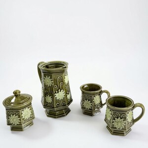 Puede incluir: Un juego de té de cerámica en color verde oliva que incluye una jarra, un azucarero con tapa y dos tazas. Cada pieza tiene una base octogonal y está decorada con motivos de margaritas en relieve de color verde claro y patrones de enredaderas.