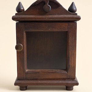 May include: A small, dark brown wooden cabinet with a square glass-front door and a round knob. The top features a curved pediment with two pointed finials on either side, and the base is supported by four small, rounded feet.