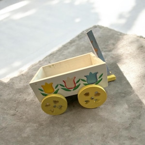 Pode incluir: Um pequeno vagão de brinquedo de madeira pintado de branco com desenhos florais amarelos, vermelhos e azuis. O vagão tem rodas amarelas e uma alça azul. É uma peça decorativa charmosa.