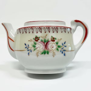 Könnte beinhalten: Eine weiße Keramik-Teekanne mit einem handgemalten Blumenmotiv aus rosa und weißen Rosen, grünen Blättern und blauen Akzenten. Der Rand ist mit einer roten Linie und einem Muster verziert, während der Griff und der Ausguss rote Details aufweisen.
