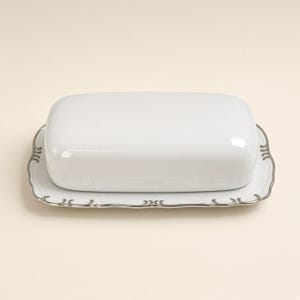 May include: A rectangular white ceramic butter dish with a smooth, domed lid. The base tray features a scalloped edge with a metallic silver trim, showcasing a decorative, repeating scroll pattern along the entire perimeter of the dish.