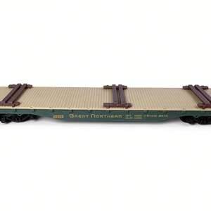 May include: A model flatcar train carriage with a green metal body and a tan wooden-textured deck. The side features the text GREAT NORTHERN, the number 42953, and technical data. Three pairs of brown wooden beams are secured across the deck.