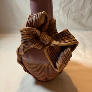 Könnte beinhalten: Eine handgefertigte Keramikvase in Mauve mit einem strukturierten, bogenförmigen Design in Brauntönen. Die Vase hat einen zylindrischen Hals und einen gerippten Boden, was einen einzigartigen künstlerischen Stil zeigt. Die Höhe beträgt ca. 20 cm.