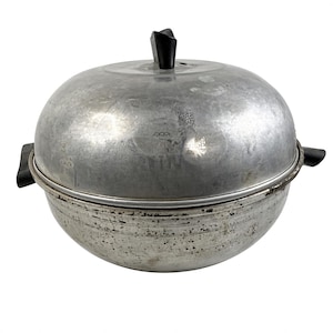 Puede incluir: Una olla redonda de metal plateado con una tapa abovedada a juego. La olla y la tapa tienen asas de plástico negro en los lados y un pomo de plástico negro en la parte superior. La superficie exterior muestra signos de desgaste y manchas.