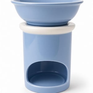May include: A light blue ceramic wax warmer featuring a cylindrical base with a large arched opening at the bottom and a removable bowl at the top. The piece includes a thick white ring detail separating the bowl from the main body of the warmer.