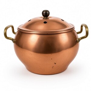 May include: A round copper pot with two brass side handles and a matching copper lid. The lid features a small dark knob handle at the center and a series of circular ventilation holes arranged in a ring around the top surface of the lid.