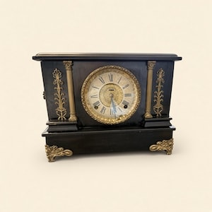 Puede incluir: Un reloj de sobremesa rectangular negro con una esfera circular central con números romanos, un fondo dorado de filigrana y adornos de metal color latón, que incluyen columnas estriadas, paneles laterales y pies tallados.