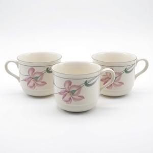 May include: Three cream-colored ceramic mugs featuring a delicate floral pattern of pink and yellow iris flowers with green stems. Each mug has a rounded handle, a smooth finish, and a thin grey line encircling the upper rim of the vessel.