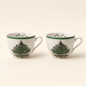 May include: Two white ceramic tea cups featuring a green rim and a detailed illustration of a decorated Christmas tree with colorful ornaments and gifts at the base. Each cup has a curved handle and a smooth, rounded body design.