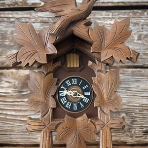 May include: A brown wooden cuckoo clock featuring a carved bird at the top and maple leaf accents surrounding the frame. The clock face is black with white Roman numerals and gold-colored hands, with the text W. GERMANY printed at the bottom.