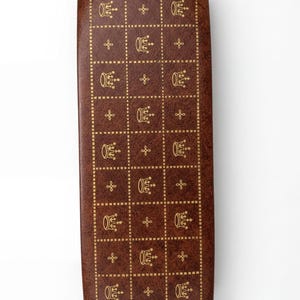 May include: A rectangular brown case with a textured surface featuring a gold grid pattern. Each square in the grid contains either a small gold crown icon or a gold floral motif, arranged in alternating rows across the entire surface.