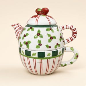 May include: A ceramic tea for one set featuring a teapot stacked on a teacup. The teapot has a white base with green holly leaves and red berries, a red and white striped handle and spout, and a lid topped with two red berries.
