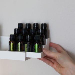 Könnte beinhalten: Ein weißes, abgestuftes Holzregal mit zwölf bernsteinfarbenen Glasflaschen für ätherische Öle mit schwarzen Verschlüssen. Jede Flasche hat ein Etikett mit dem Markennamen dōTERRA und der jeweiligen Ölsorte, darunter Bergamot, Petitgrain, Lemongrass und Eucalyptus.
