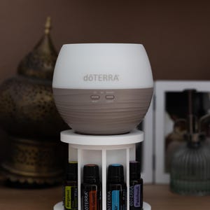 Può includere: Un diffusore di oli essenziali dōTERRA bianco e grigio è posizionato su un supporto circolare bianco a più livelli. Il supporto presenta quattro scomparti che contengono piccole boccette di vetro scuro con etichette Bergamot, On Guard, Air e Patchouli.