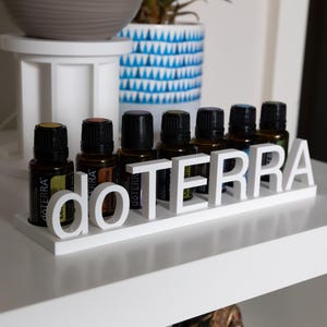 Könnte beinhalten: Ein weißer Holzständer mit dem Schriftzug dōTERRA in erhabenen Buchstaben, der Platz für sieben kleine ätherische Ölflaschen aus Braunglas mit schwarzen Verschlüssen bietet. Der Ständer steht auf einer ebenen, weißen Oberfläche.