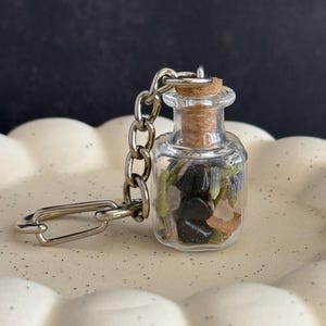 May include: A small, clear glass bottle keychain filled with various stones and dried herbs. The bottle is sealed with a cork and attached to a silver chain and keyring. The contents include black stones, lavender, and pink crystals.
