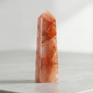 Könnte beinhalten: Ein hoher, facettierter Kristallobelisk in Orange- und Pfirsichtönen. Der polierte Edelstein hat eine spitze Spitze und eine rechteckige Basis. Der Kristall ist durchscheinend, so dass Licht hindurchscheinen kann, und ist vor einem neutralen Hintergrund platziert.