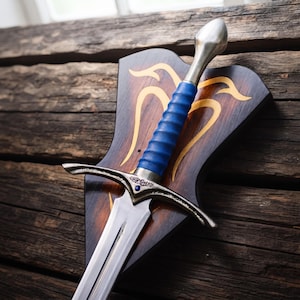 Può includere: Una spada in metallo color argento con un'impugnatura blu brillante e una piccola pietra blu incastonata nella guardia. La spada è montata su una placca di legno marrone scuro con un motivo intarsiato simmetrico e curvo di colore chiaro.