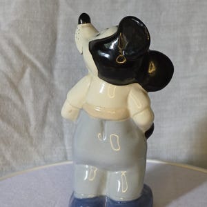 May include: A ceramic figurine of a cartoon mouse, featuring a black head and ears, a white face, and a light blue outfit. The figurine is standing on a white surface, with a light gray background.