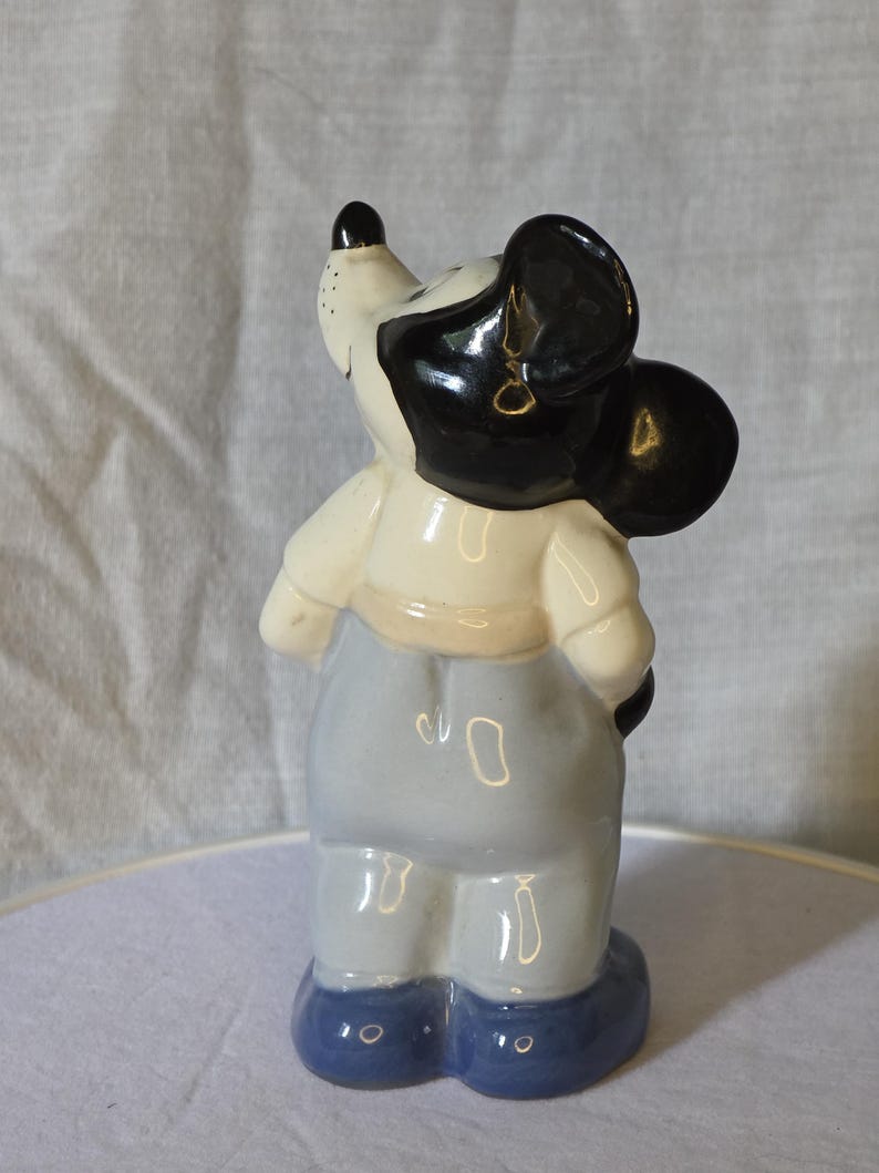 May include: A ceramic figurine of a cartoon mouse, featuring a black head and ears, a white face, and a light blue outfit. The figurine is standing on a white surface, with a light gray background.
