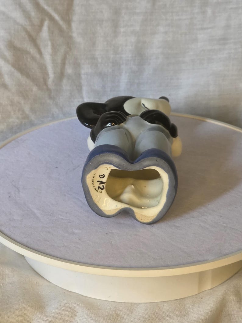May include: A vintage Mickey Mouse figurine, featuring a black head and ears, white face, and blue overalls. The figurine is ceramic and has a glossy finish. The base is marked with "D12".