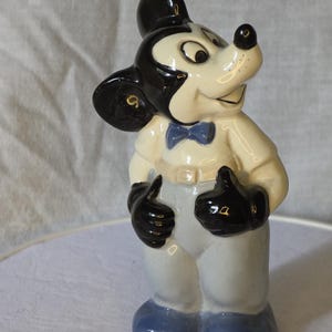 May include: A ceramic figurine of Mickey Mouse, featuring a black head and ears, a white face, and a black nose. He wears a white shirt, light blue pants, black gloves, and blue shoes. A blue bow tie completes the look.