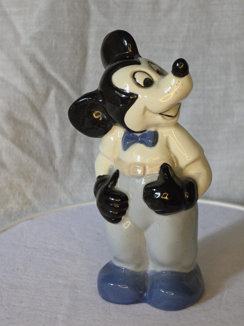 May include: A ceramic figurine of Mickey Mouse, featuring a black head and ears, a white face, and a black nose. He wears a white shirt, light blue pants, black gloves, and blue shoes. A blue bow tie completes the look.