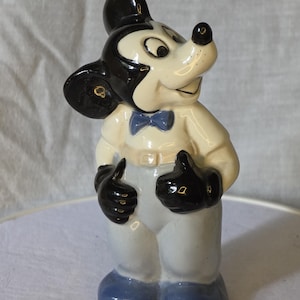 May include: A ceramic Mickey Mouse figurine. The figure has a black head and ears, a white face, and a black nose. It wears a white shirt, light blue pants, a blue bow tie, and black gloves and shoes. The figurine is standing and giving a thumbs-up.