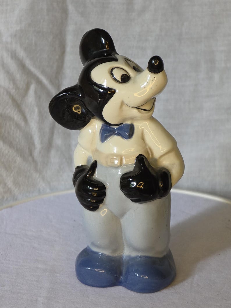 May include: A ceramic Mickey Mouse figurine. The figure has a black head and ears, a white face, and a black nose. It wears a white shirt, light blue pants, a blue bow tie, and black gloves and shoes. The figurine is standing and giving a thumbs-up.