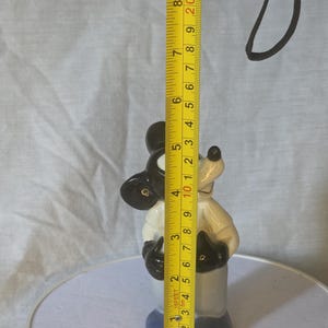 May include: A ceramic Mickey Mouse figurine, primarily black, white, and blue, stands approximately 4 inches tall. The figurine is measured with a yellow tape measure, showing its height. The background is a light gray fabric.