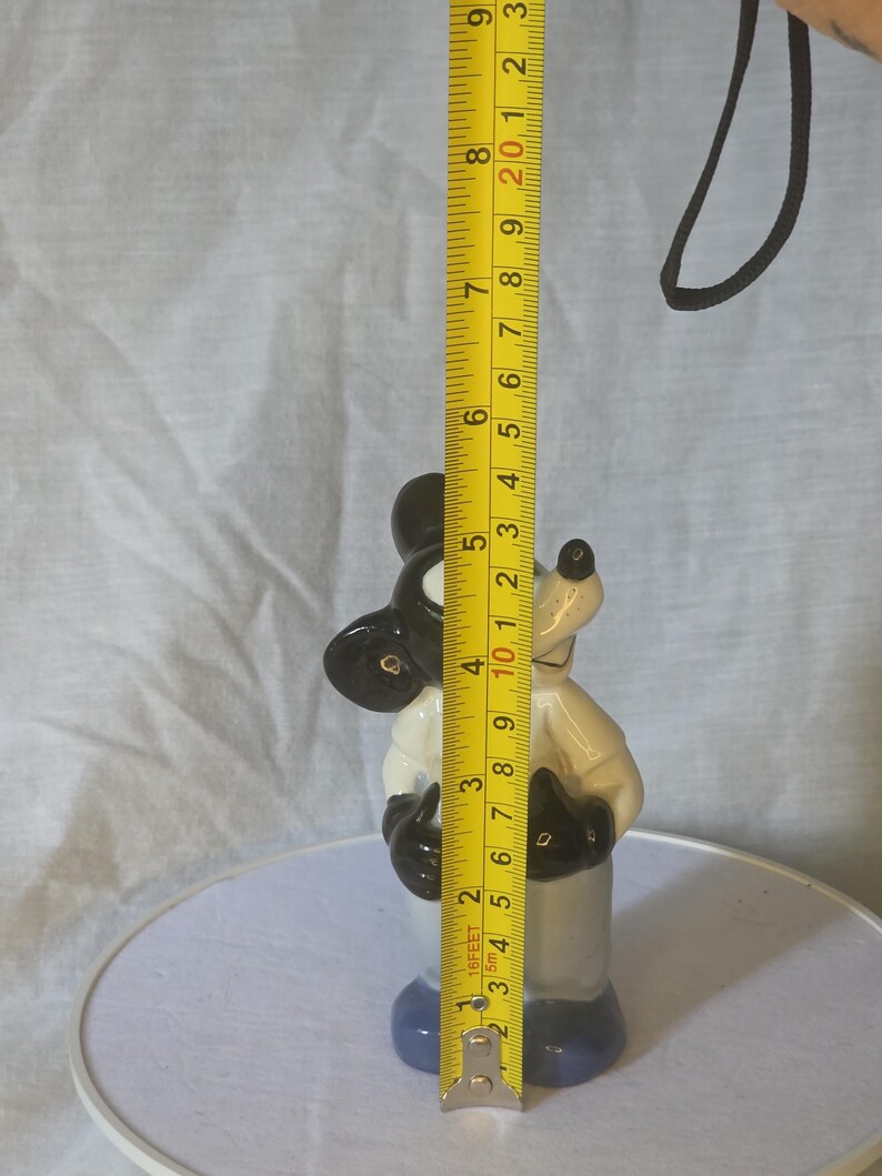 May include: A ceramic Mickey Mouse figurine, primarily black, white, and blue, stands approximately 4 inches tall. The figurine is measured with a yellow tape measure, showing its height. The background is a light gray fabric.