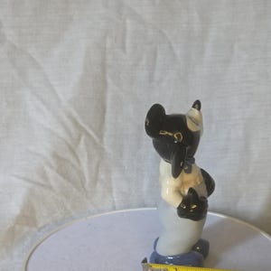 May include: A ceramic figurine of a black and white animal, possibly a cat or dog, holding a smaller black animal. The figurine has a light blue base and is approximately 3 inches tall.