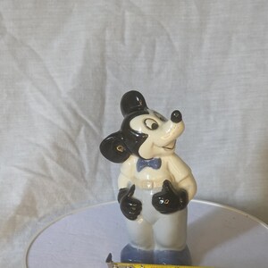 May include: A ceramic figurine of Mickey Mouse, wearing a white jacket, gray pants, and a blue bow tie. The figure has black ears, gloves, and a black hat. The figurine is approximately 4 inches tall.