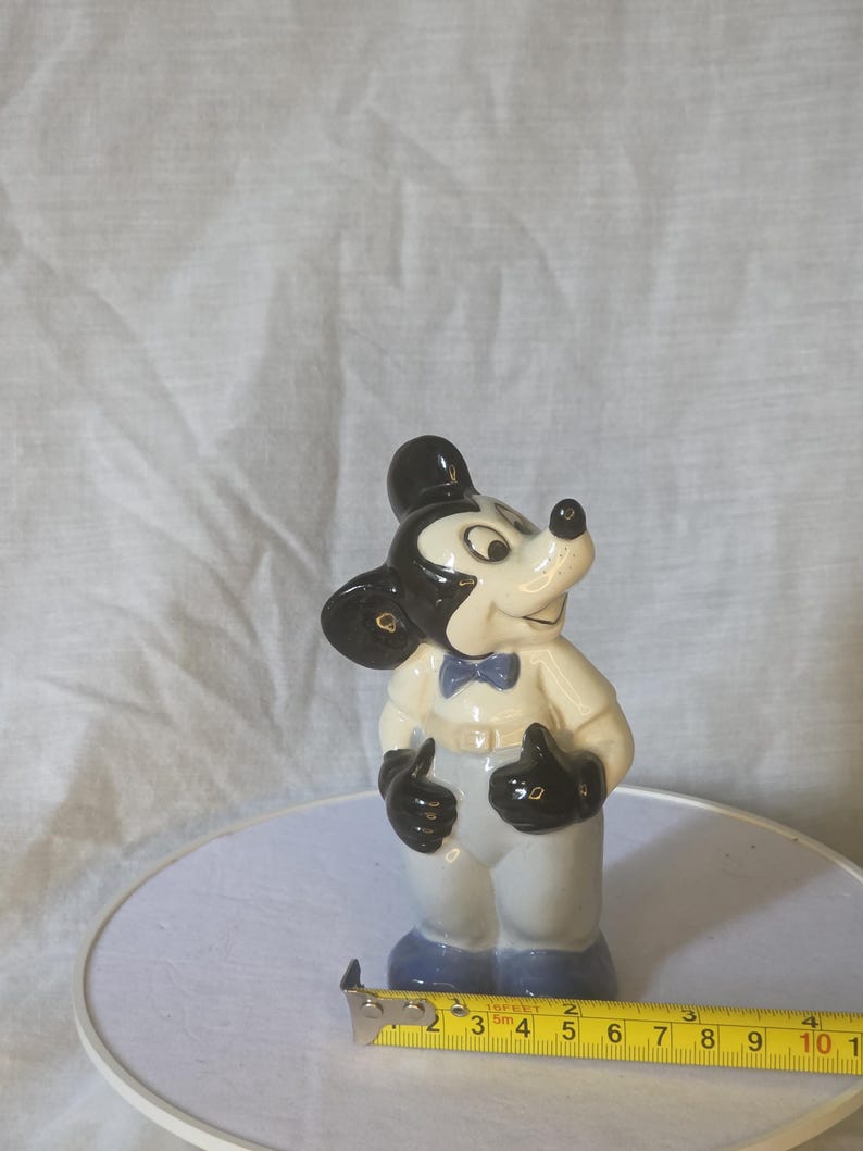 May include: A ceramic figurine of Mickey Mouse, wearing a white jacket, gray pants, and a blue bow tie. The figure has black ears, gloves, and a black hat. The figurine is approximately 4 inches tall.