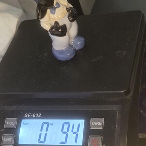 May include: A vintage ceramic figurine of a cartoon character, featuring black, white, and blue accents. The figurine is standing on a black electronic scale, which displays a weight of 0.94 kg.