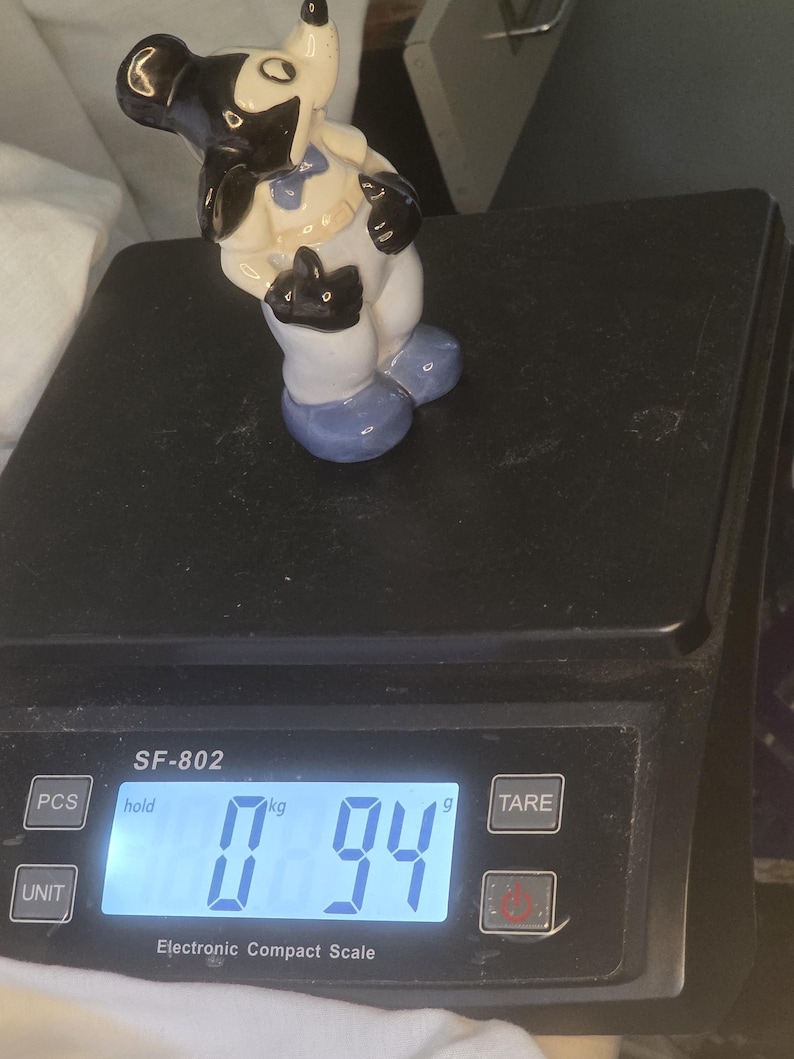 May include: A vintage ceramic figurine of a cartoon character, featuring black, white, and blue accents. The figurine is standing on a black electronic scale, which displays a weight of 0.94 kg.