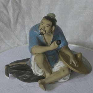 Può includere: Statuetta in ceramica di un uomo seduto con una camicia blu e pantaloni bianchi. Ha la barba e tiene in mano un piccolo oggetto nero. Un cappello a tesa larga è appoggiato accanto a lui. La statuetta è su un cuscino nero.