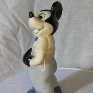 May include: A ceramic figurine of Mickey Mouse, featuring black ears, a black nose, and a smiling expression. The figure wears a light blue outfit with black gloves and blue shoes. The figurine is a collectible item.