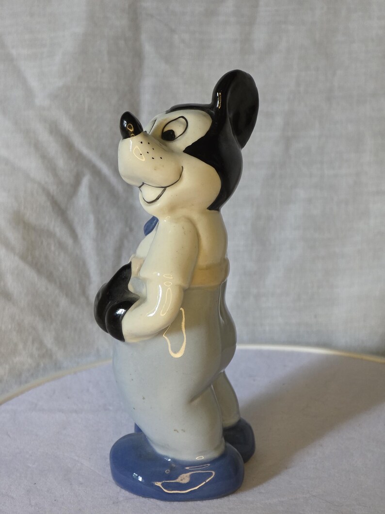 May include: A ceramic figurine of Mickey Mouse, featuring black ears, a black nose, and a smiling expression. The figure wears a light blue outfit with black gloves and blue shoes. The figurine is a collectible item.