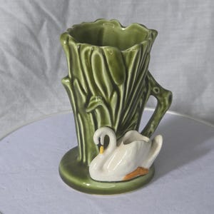 Peut inclure: Un vase en céramique vert avec un motif de tronc d'arbre et une figurine de cygne blanc. Le vase a une surface texturée et une poignée. Le cygne a des pieds orange et un bec jaune. Le vase est sur une base ronde.