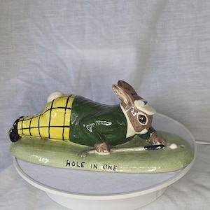 Op de afbeelding: Een keramieken konijnfiguur in een groene jas en een gele geruite broek, golf aan het spelen. Het beeldje staat op een groene basis met de tekst "HOLE IN ONE". Het konijn is bruin en wit.
