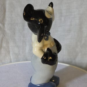 May include: A ceramic figurine of a cartoon character with a black head and ears, wearing a light blue outfit with a white shirt and black gloves. The figurine has a glossy finish and is standing on a blue base.