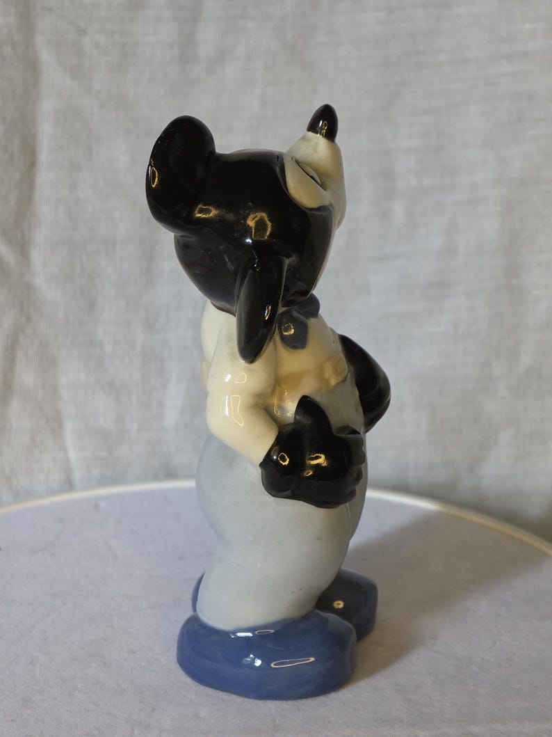 May include: A ceramic figurine of a cartoon character with a black head and ears, wearing a light blue outfit with a white shirt and black gloves. The figurine has a glossy finish and is standing on a blue base.