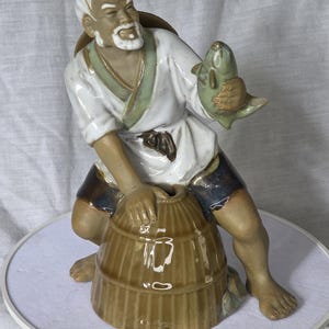 May include: A ceramic figurine of an elderly fisherman in a white and green robe, holding a fish. The figurine is kneeling beside a brown basket. The figure has a white beard and is wearing dark blue shorts. The figurine is on a white surface.