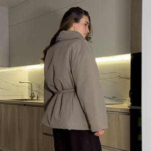 Könnte beinhalten: Eine taupefarbene Steppjacke mit passendem Bindegürtel, langen Ärmeln und hohem Kragen, kombiniert mit einer locker sitzenden schwarzen Hose und schwarzen Schlupf-Stiefeletten mit dicker Sohle, von hinten und der Seite gesehen.