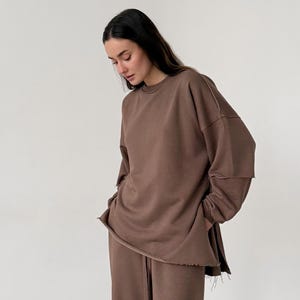 Puede incluir: Un conjunto marrón de sudadera y pantalón ancho a juego. La sudadera tiene cuello redondo y detalles de dobladillo sin rematar. El pantalón tiene cintura con cordón. El conjunto se combina con zapatillas blancas. El conjunto de ropa es de material suave.