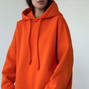 May include: A bright orange hooded sweatshirt with long sleeves, ribbed cuffs, and a matching drawstring at the neck. The garment features a relaxed fit with dropped shoulders and a straight hemline, crafted from a solid-coloured fabric.
