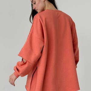 Puede incluir: Una sudadera de color coral de gran tamaño con un corte holgado y mangas largas. La sudadera presenta un diseño de manga dividida y un dobladillo dividido. La tela parece ser un material suave y cómodo, adecuado para ropa informal.